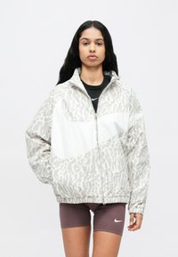 Επένδυση με φερμουάρ σε ανοιχτό γκρι χρώμα με διακριτικό σχέδιο λεοπάρ, με λευκή επιφάνεια και ελαστικές μανσέτες. Συνδυάζεται με σκούρες σορτς.