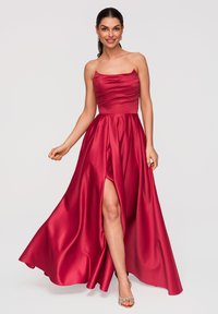Femme en robe rouge en satin sans bretelles et longue jusqu'au sol, avec une fente à l’avant, souriante et posant avec une main légèrement levée.