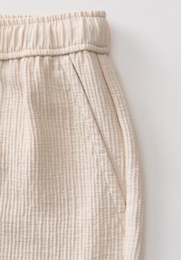 Pantalon texturé beige avec ceinture élastique et poche latérale inclinée, présenté en gros plan sur fond blanc.