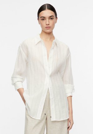 Kobieta w białej teksturowanej koszuli zapinanej na guziki z podwiniętymi rękawami i beżowych spodniach, stojąca na jednolitym białym tle.