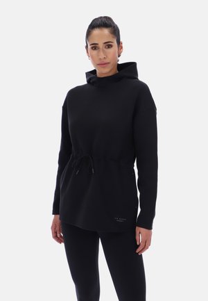 Kvinde med trukket hår tilbage, iført en sort langærmet hoodie med snor i taljen og sorte leggings.