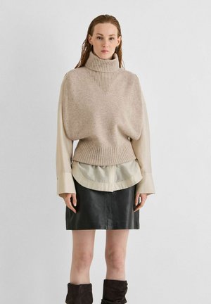Mujer con suéter de cuello alto beige sobre camisa crema, falda de cuero negra y botas negras holgadas, de pie frente a un fondo blanco liso.