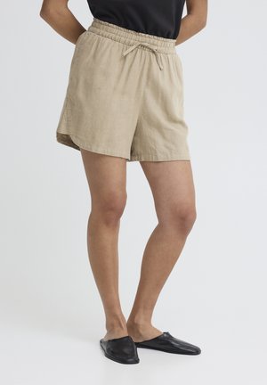 Osoba v béžových ľanových kraťasoch so šnúrkou a čiernych nazúvacích topánkach, stojaca s rukami v vreckách pred obyčajným pozadím.