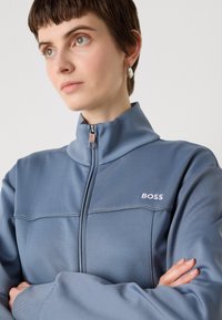 Light blue zip-up jacket with high collar and white embroidered "BOSS" logo. Smooth fabric, relaxed fit, and subtle stitching details.
