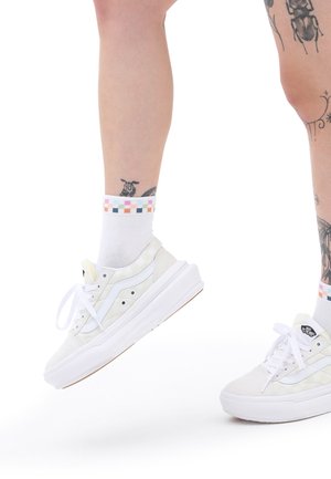 White canvas sneakers with white accents and rubber sole, paired with multicolored checkered ankle socks. Visible tattoos on the leg.