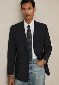 Blazer bleumarin cu două rânduri de nasturi, dintr-un material texturat, având rever larg și două buzunare frontale, asortat cu o cravată cu model și blugi din denim deschis.