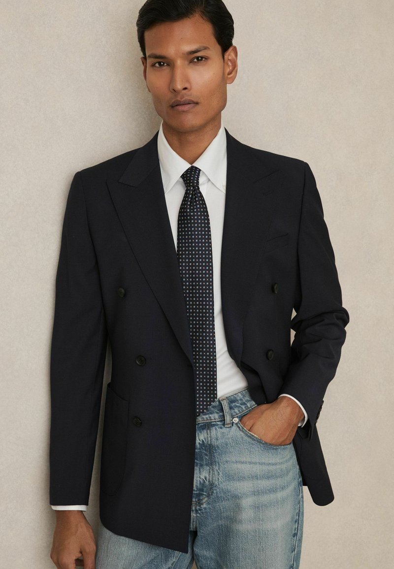 Blazer bleumarin cu două rânduri de nasturi, dintr-un material texturat, având rever larg și două buzunare frontale, asortat cu o cravată cu model și blugi din denim deschis.