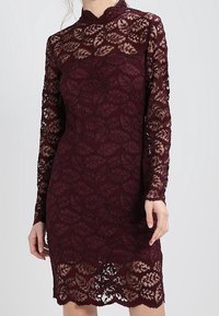 Burgundské krajkové šaty s dlouhými rukávy a vysokým výstřihem, zdobené vzorem listů a zvlněným lemem. Mírně průhledná textura.