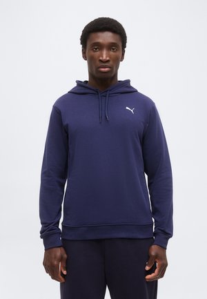 Junger Mann, der nach vorne blickt und einen marineblauen Puma-Hoodie mit weißem Logo auf einem einfarbigen hellen Hintergrund trägt.