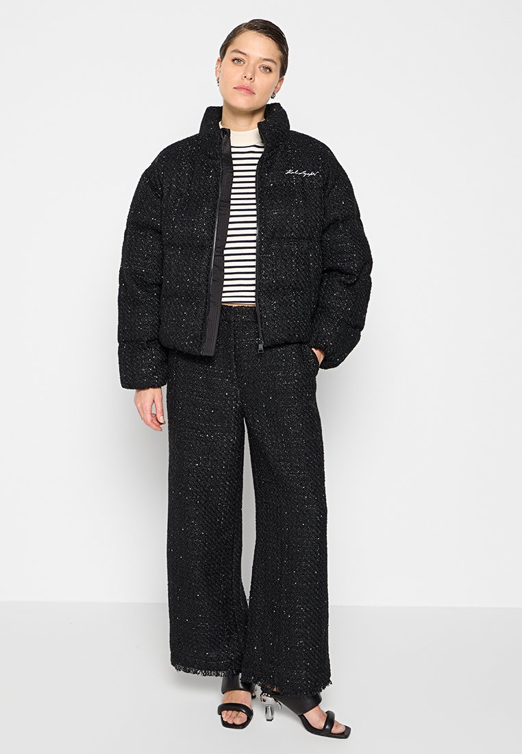 Schwarze Daunenjacke mit gestepptem Design, kombiniert mit schwarzen weit geschnittenen Hosen mit einem fransigen Saum. Darunter ist ein gestreiftes Oberteil sichtbar.