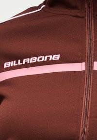 Κοντινό πλάνο καφέ μπουφάν με φερμουάρ, με ροζ οριζόντια γραμμή και λογότυπο "BILLABONG" στο στήθος.