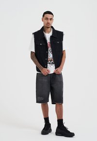 Vest in denim nero sopra una t-shirt bianca con grafica colorata. Pantaloni corto in denim grigio con dettagli sfilacciati e sneaker nere.