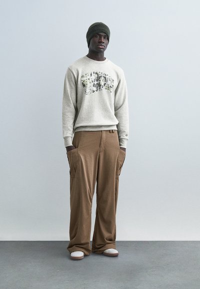 Mann iført grå genser med grafisk trykk, brune vide bukser med lommer, grønn strikket lue og hvite joggesko, stående mot ensfarget bakgrunn.