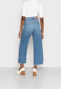 Džíny z modrého denimu s vysokým pasem a širokými nohavicemi, s klasickým pětizásuvkovým designem a třepeným lemem. Kombinují se s krémovými botami na blokovém podpatku.