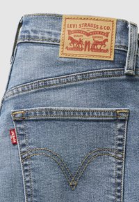 Világoskék farmernadrág barna bőr címkével, piros varrással és jellegzetes V alakú hátsó zsebvarrással.