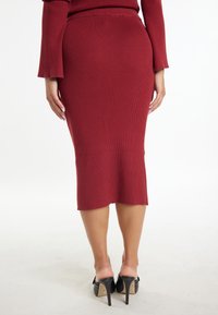 Jupe crayon en tricot bordeaux avec une texture côtelée, longueur genoux, dotée d'une légère fente à l'ourlet. Portée avec des chaussures à talons noirs.