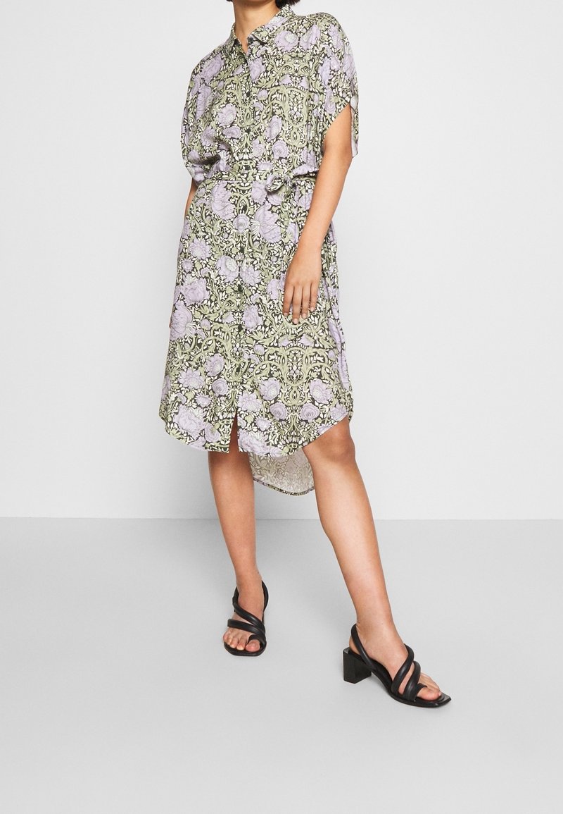 Robe courte à manches courtes, fleurie en violet et vert, avec col, fentes latérales et tissu léger ; associée à des sandales noires à talons et brides.