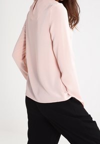 Pink long-sleeve blouse with a back neckline split, made of lightweight, textured fabric, paired with black trousers.