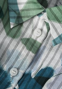Primo piano di una camicia a righe con motivi astratti verdi, blu e bianchi, caratterizzata da bottoni bianchi e colletto piegato.