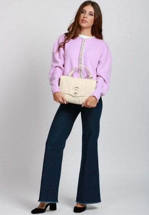 Donna che indossa un maglione lilla con dettagli in perla, jeans scuri a zampa, tacchi neri, che tiene una borsa beige su uno sfondo chiaro uniforme.