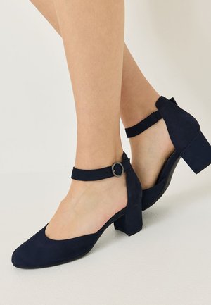 Donna che indossa scarpe in camoscio blu navy con tacco a blocchetto, cinturini alla caviglia e fibbie tonde argentate, in posa su sfondo neutro.
