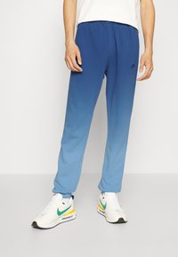 Modré gradientové tepláky s elastickým pasem a manžetami, vyrobené z měkké tkaniny. Nosené s bílými teniskami s barevnými akcenty.