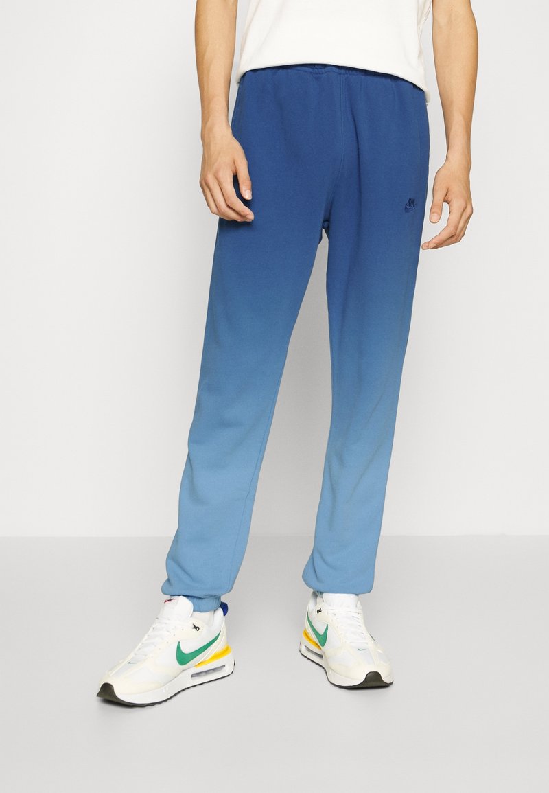 Modré gradientové tepláky s elastickým pasem a manžetami, vyrobené z měkké tkaniny. Nosené s bílými teniskami s barevnými akcenty.