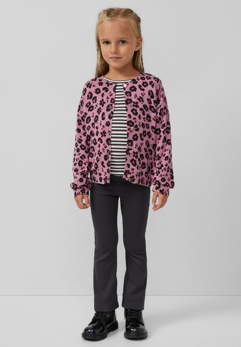 Rosa Leopardenmuster-Cardigan über einem schwarz-weiß gestreiften Hemd, kombiniert mit dunkelgrauen Hosen und glänzenden schwarzen Stiefeln.
