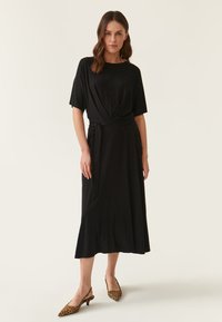 Schwarzes Midi-Kleid mit kurzen Ärmeln, Wickeldesign und gebundenem Taillengürtel. Hat eine glatte Textur und fließende Silhouette. Tiermuster-Schuhe.