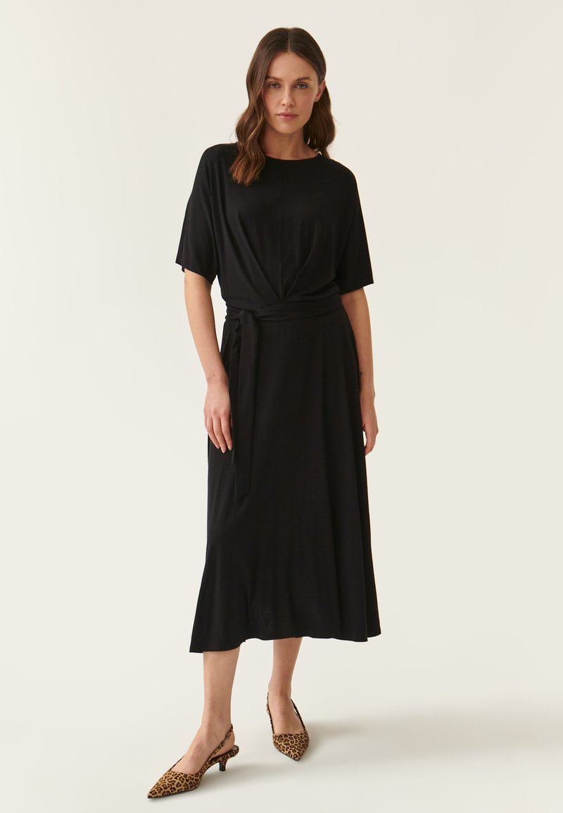Schwarzes Midi-Kleid mit kurzen Ärmeln, Wickeldesign und gebundenem Taillengürtel. Hat eine glatte Textur und fließende Silhouette. Tiermuster-Schuhe.