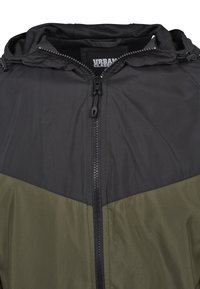 Giacca con cappuccio e chiusura a zip, caratterizzata da una parte superiore nera e una parte inferiore verde oliva, realizzata in un tessuto liscio e resistente.