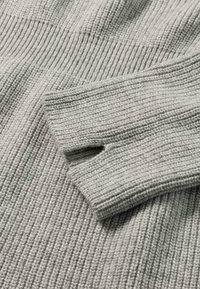 Maglione lavorato a maglia grigio chiaro con texture a coste e design delle maniche ripiegate, caratterizzato da una leggera variazione nel motivo di lavorazione.