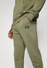 Olīvu zaļš kapuci jaku un sporta bikses komplekts, izgatavots no mīksta materiāla. Iespējamie melni zīmola etiķetes priekšpusē, ar brīvu piegriezumu un sānu kabatām.