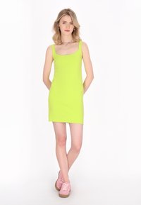 Rochie fără mâneci de culoare verde neon, cu decolteu pătrat și siluetă ajustată. Modelul poartă tenisi roz cu tălpi maro, stând în fața unui fundal alb.