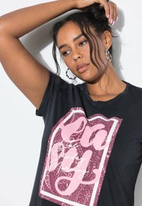 Camiseta de algodón negra con un gran gráfico rosa que presenta patrones intrincados y texto blanco. Mangas cortas y cuello redondo.