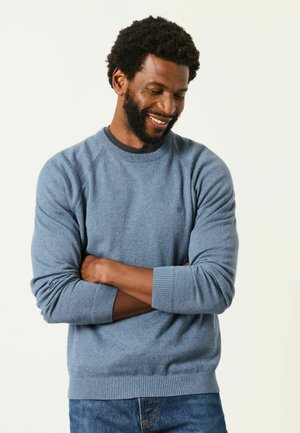 Mann mit Bart und lockigem Haar, trägt einen blauen Pullover und Jeans, lächelt und schaut nach unten mit verschränkten Armen.