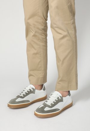 Hvide og grå sneakers med ruskind- og mesh-teksturer, der har en rund tå, snøre fortil og gummigummi sål, båret med beige bukser.