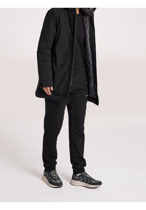 Giacca nera isolata con un rivestimento interno trapuntato, abbinata a pantaloni neri e sneakers scure. La giacca presenta chiusura a zip e bottone.