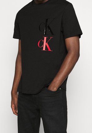 Mann trägt schwarzes T-Shirt mit schwarzem und rotem "cK"-Logo auf der Brust, kombiniert mit schwarzer Jeans, steht mit einer Hand in der Tasche.