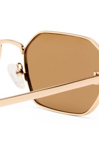 Κοντινή λήψη χρυσών εξαγωνικών γυαλιών ηλίου με καφέ σκουρόχρωμους φακούς και διαφανείς ρυθμιζόμενες μύτες στη λευκή επιφάνεια.