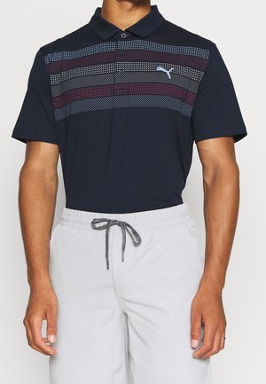 Homme portant un polo bleu marine à rayures horizontales rouges et blanches en points, et un pantalon gris clair avec cordon de serrage.