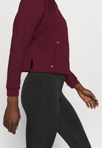 Persona che indossa un top a maniche lunghe bordeaux con spacchi laterali e leggings neri aderenti, in piedi di fronte a uno sfondo chiaro e uniforme.