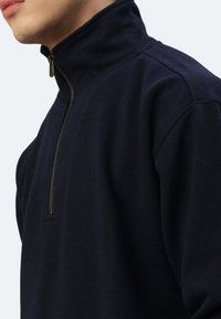 Marineblauer Reißverschluss-Sweatshirt mit hohem Kragen, ausgestattet mit einem bronzefarbenen Reißverschluss und weichem, strukturiertem Stoff. Das Design beinhaltet Schulternähte und eingesetzte Ärmel.