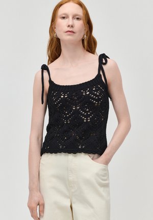 Nő hosszú vörös hajjal, fekete horgolt pánt nélküli felsőt visel, melynek pántjai megkötve, és világos bézs nadrágot, egyik kezét zsebre téve.