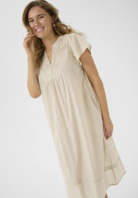 Robe beige à manches courtes avec un décolleté en V, corps plissé et couture décorative à l'ourlet. Fabriquée en tissu léger.