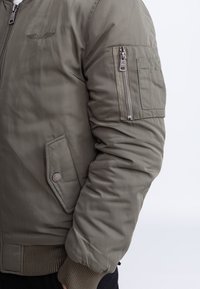 Primer plano de persona con chaqueta bomber verde oliva con bolsillo con cremallera en el brazo, bolsillo lateral con solapa, puños acanalados y logo de ala bordado en el pecho.