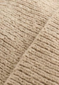 Tissu beige tricoté avec des nervures texturées, présentant des fibres douces et duveteuses. Le matériau semble chaud et confortable, adapté pour des vêtements ou de la décoration.