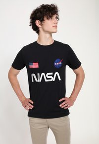 �Černé bavlněné tričko s nápisem "NASA" ve velkých bílých písmenech, nášivkou americké vlajky na levém rukávu a logem NASA na hrudi.