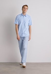 Camisa de manga corta azul claro con un bolsillo en el pecho, combinada con jeans azul claro. Los zapatos son blancos con suela marrón. Diseño sencillo y limpio.