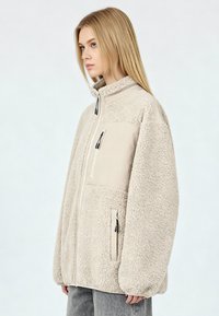 Giacca in pile beige con collo alto, tasche con cerniera e pannello anteriore liscio. Presenta una superficie morbida e texturizzata e una vestibilità comoda.